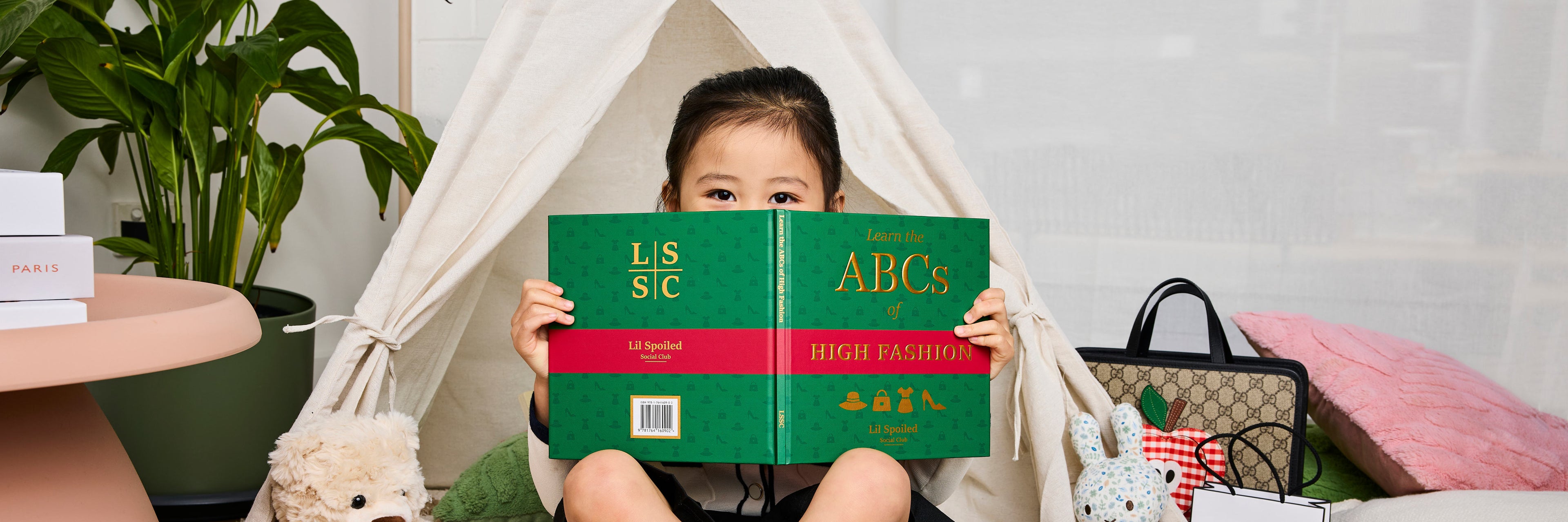 Child holding a book titled 'Learn the ABCs of High Fashion' in a cozy indoor setting with a plant and furniture.
