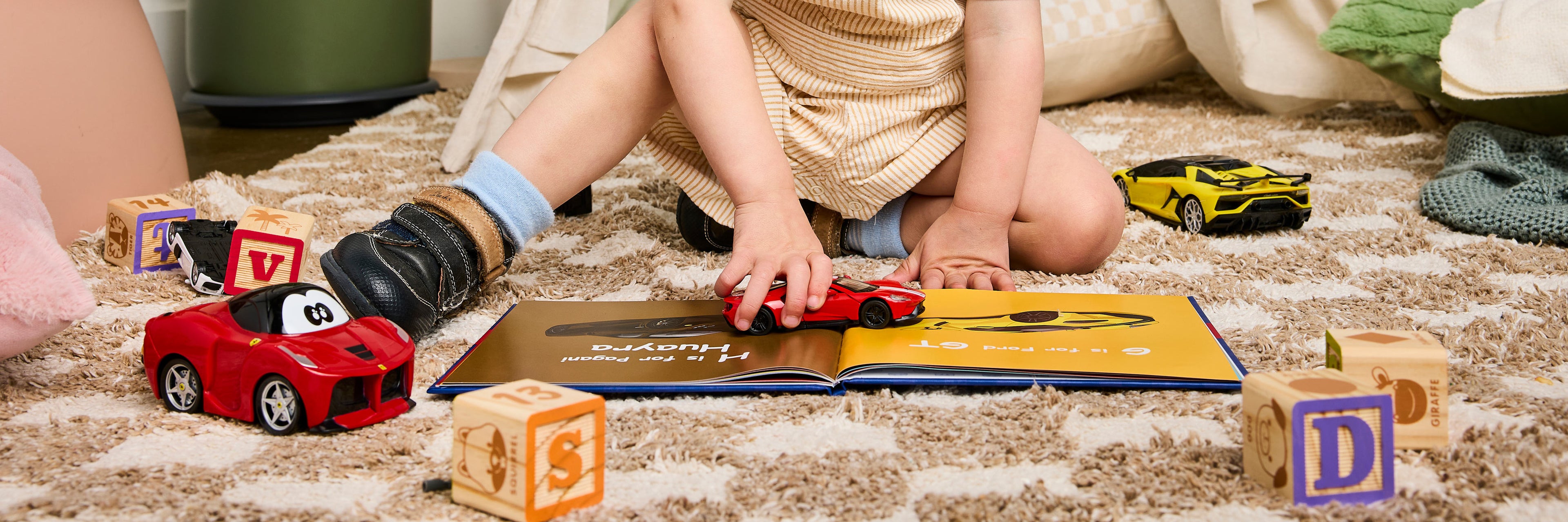 Child playing with toy cars and blocks on a carpeted floor. 'Learn the ABCs of Supercars' is a book on the floor.
