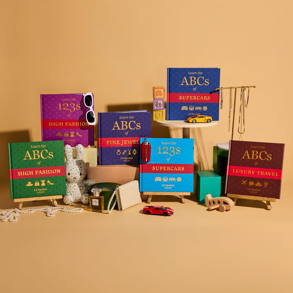 A styled product display of miniature book-shaped accessories and toys on a warm beige background. The set includes colorful mini books titled 'Learn the ABCs of [topic]' on small easels, along with small accessories like a plush bunny, sunglasses, a toy car, a miniature suitcase, and jewelry stands.