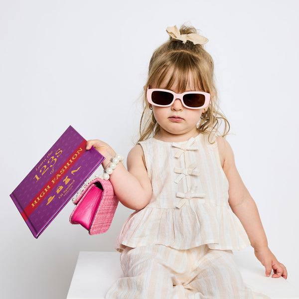 Child in a white dress with pink sunglasses holding a '123s of High Fashion' book and pink shoes on a white background