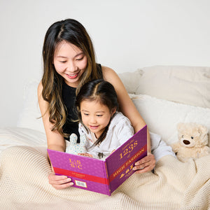 Woman/parent and child reading a book together on a bed with a teddy bear. The book is 'Learn the ABCs of High Fashion'.