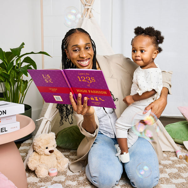 Woman reading a 'Learn the 123s of High Fashion' book to a child in a cozy living room setting.