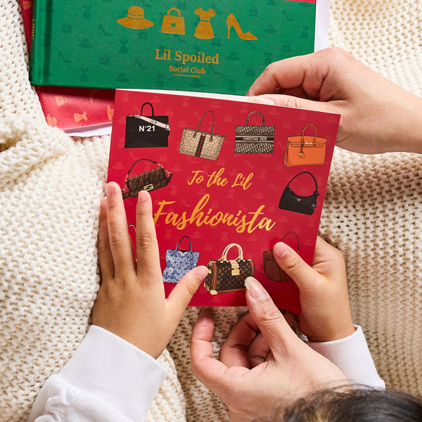 Two hands holding a red card with handbag illustrations and text, with a green book labeled 'Lil Spoiled Social Club' in the background.