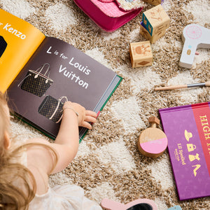 Child reading a book titled 'L is for Louis Vuitton' on a carpeted floor with toys around.