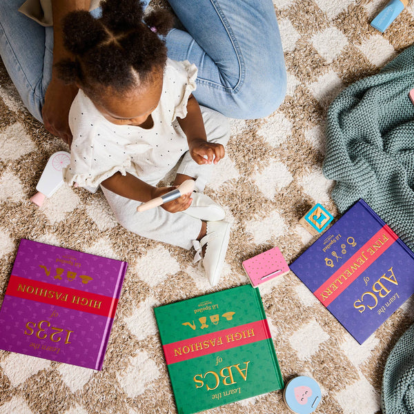 Child sitting on a carpeted floor with a makeup set and educational ABCs and 123s books and toys.